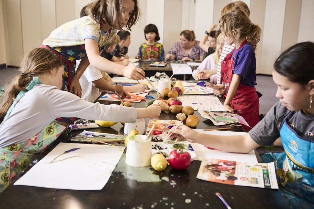 Primary school students painting a still life of fruit at a long table during an art class